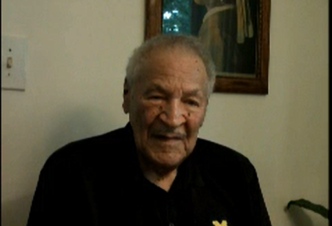Black man in his late 80s sitting for an interview; he wears a navy polo shirt with the University of Michigan logo embroidered on the chest.